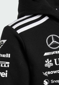Sudadera negra con franjas horizontales blancas en los hombros, con varios logotipos en texto blanco, incluyendo un emblema de Mercedes-Benz.
