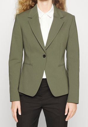 Blazer ajusté vert olive avec un col cranté, fermeture à un bouton et poche frontale unique, associé à une chemise blanche en dessous.