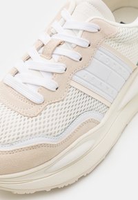 Ljusbeige och vit mesh-sneaker med mockadetaljer, med vita snören och en texturerad sula. "TOMMY JEANS"-logotyp på sidan.