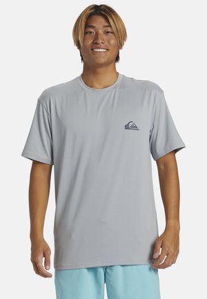 Junger Mann lächelt, trägt ein hellgraues kurzärmeliges T-Shirt mit einem kleinen dunklen Logo auf der Brust und hellblaue Shorts.