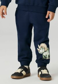 Barn iført marineblå joggingbukser med en triceratops-dinosaurgrafik på den nederste del af benet og sorte sneakers med hvide snørebånd.