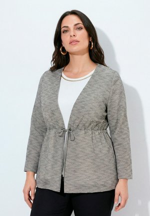 Femme aux cheveux foncés portant des boucles d'oreilles anneaux en or, un haut blanc avec un col en perles dorées et une veste grise rayée nouée à la taille.