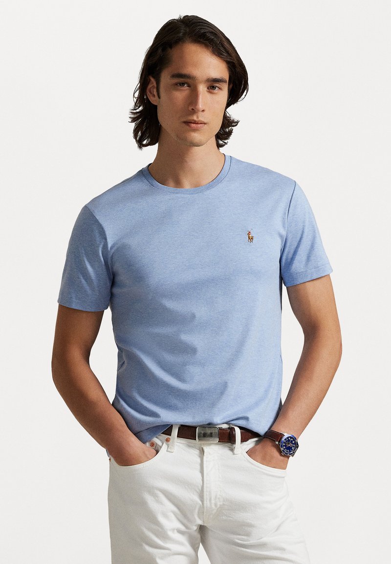 Gaiši zils kokvilnas T-krekls ar apaļu kakla un īsām piedurknēm, ar mazu izšūtu logotipu uz krūtīm. Valkāts ar baltām biksēm.