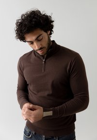 Brauner Zip-Hoodie mit langen Ärmeln, feiner Strickstruktur und kontrastierenden beigefarbenen Akzenten an den Ärmeln. Model trägt Blue Jeans.
