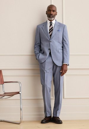 Man with gray beard wearing light blue checked suit, striped tie, and brown shoes, standing next to a modern brown leather chair.