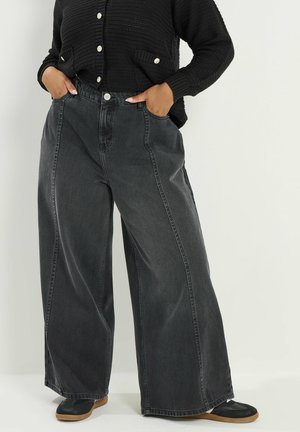 Person trägt weit geschnittene, ausgebleichte schwarze Jeans, schwarze Strickjacke mit goldenen Knöpfen, schwarze Schuhe mit hellbraunen Sohlen, Hände in den Taschen.