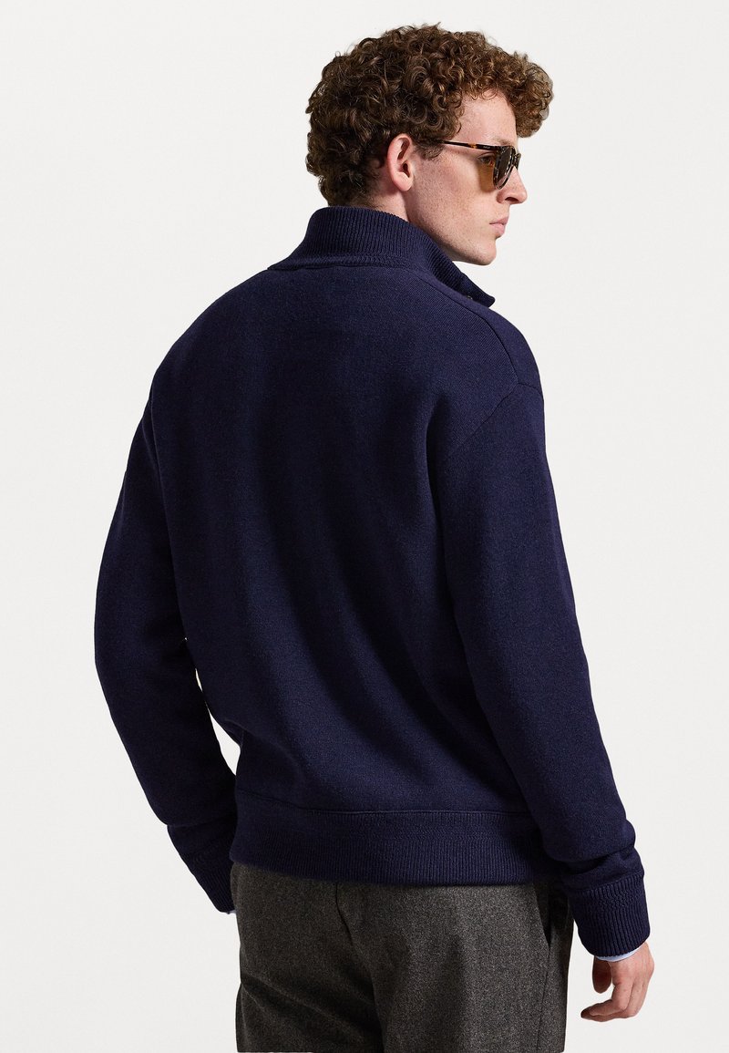 Temno modra pletena jopica z visokim ovratnikom, rebrastimi manšetami in robom. Gladka struktura, prilagojena silhueta. Obuta z sivimi hlačami.