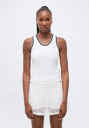 Femeie purtând un top sportiv fără mâneci, alb cu margine neagră, și o fustă albă cu model de dantelă, stând în fața unui fundal simplu și luminos.