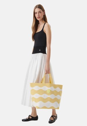 Donna con canotta nera, gonna midi bianca e ballerine nere che tiene una grande borsa a tracolla gialla e bianca con motivo ondulato.