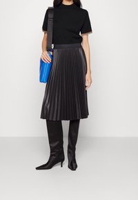 Schwarzer, plissierter Lederrock, kombiniert mit einem schwarzen Oberteil mit mehrfarbiger Borte, einer blauen Tasche und knielangen, schwarzen spitzen Stiefeln.