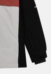 Veste colorée en noir, blanc et rouille. Dotée d'un tissu texturé, de poignets ajustables avec Velcro et de poches discrètes.