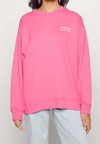 Kvinna iklädd en ljusrosa Hollister California sweatshirt tillsammans med ljusblå jeans och rosa örhängen, stående mot en enfärgad bakgrund.