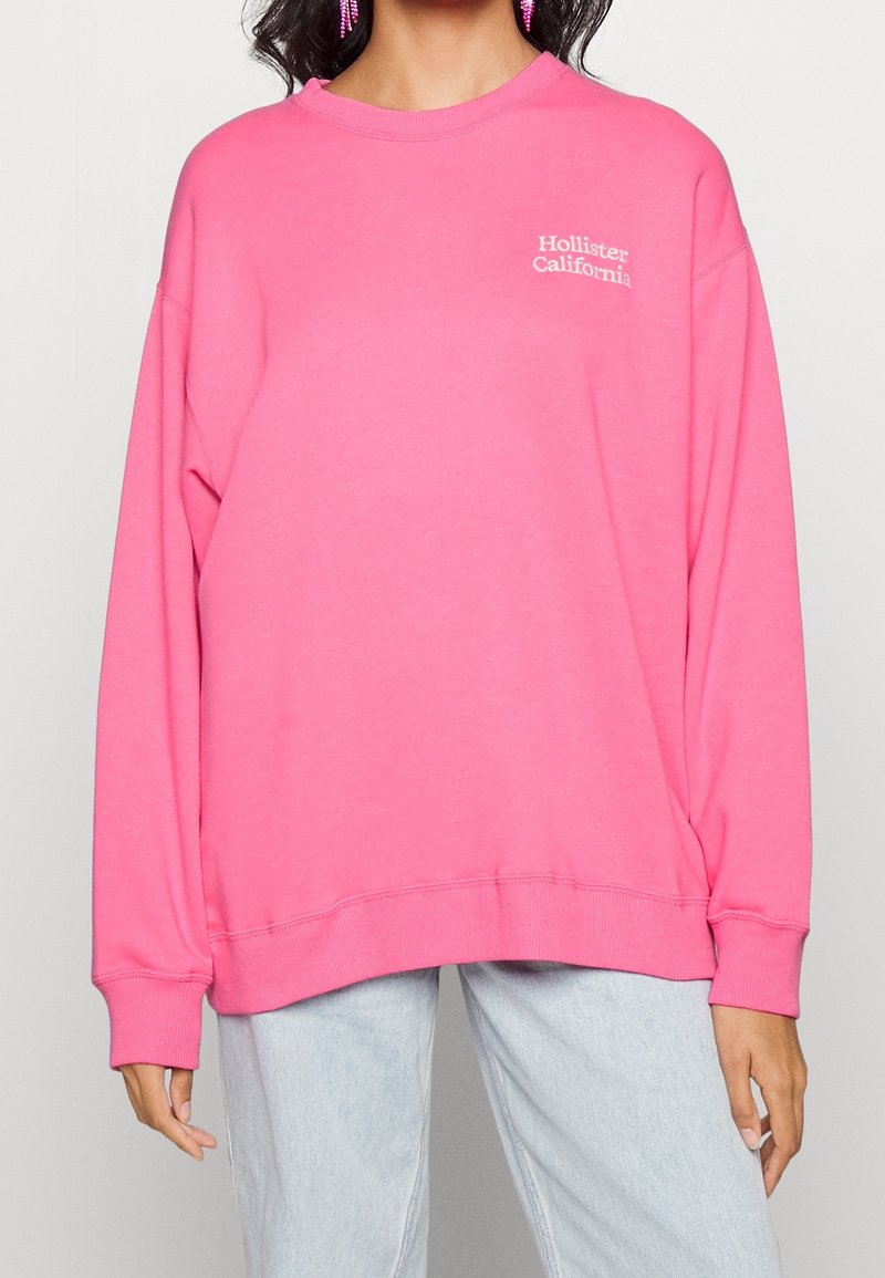 Kvinna iklädd en ljusrosa Hollister California sweatshirt tillsammans med ljusblå jeans och rosa örhängen, stående mot en enfärgad bakgrund.