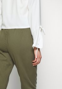 Camicia bianca con maniche a campana caratterizzata da dettagli con laccetti, abbinata a pantaloni verdi oliva; tessuto morbido e vestibilità rilassata.