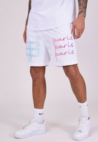 Pantalones cortos deportivos blancos con el texto "Project" y "Paris" en azul y rosa, con una textura suave y un corte relajado. Combinados con zapatillas blancas.