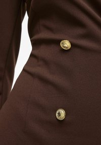 Κοντινή λήψη καφέ υφάσματος με δύο λεπτομερείς χρυσούς κουμπιά με χαραγμένο σχέδιο, τοποθετημένα κάθετα σε ένδυμα.