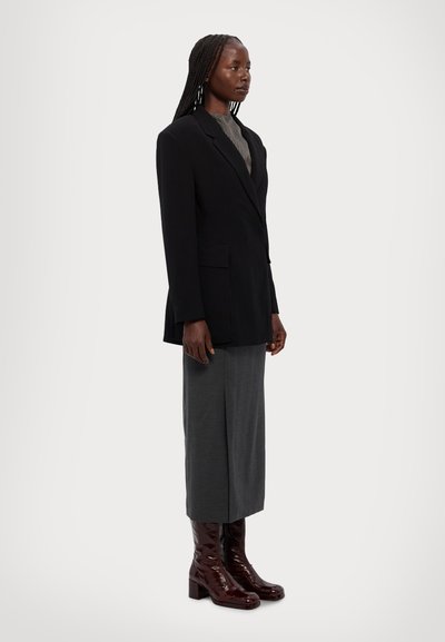 Femeie cu cosițe lungi, îmbrăcată într-un sacou negru supradimensionat, fustă midi gri, bluză cu model și cizme cu toc maro închis lucioase, stând de-o parte.