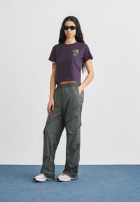 Μοβ κοντομάνικη μπλούζα με γραφικό τύπωμα στο στήθος, συνδυασμένη με χαλαρή, ελιάς πράσινη παντελόνα cargo με πολλές τσέπες.