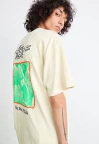Beige oversized t-skjorte laget av bomull, med et grønt grafisk rektangel omkranset av oransje søm med "Big Bend, TEXAS" trykt under.
