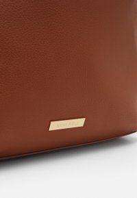 Hoek van een bruin leren tas met een klein rechthoekig gouden metalen label dat "ANNA FIELD" is gegraveerd, op een witte ondergrond.