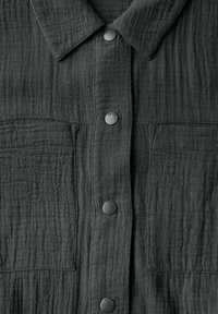 Camicia grigia scura con un tessuto testurizzato e spiegazzato, chiusure a bottone e due tasche frontali. I bottoni sono rotondi e piatti con una finitura opaca.