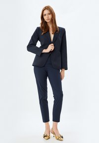 Blazer navy a righe sottili con un taglio sartoriale, abbinato a pantaloni navy slim e scarpe dorate metallizzate decorate con un accento a fiocco.