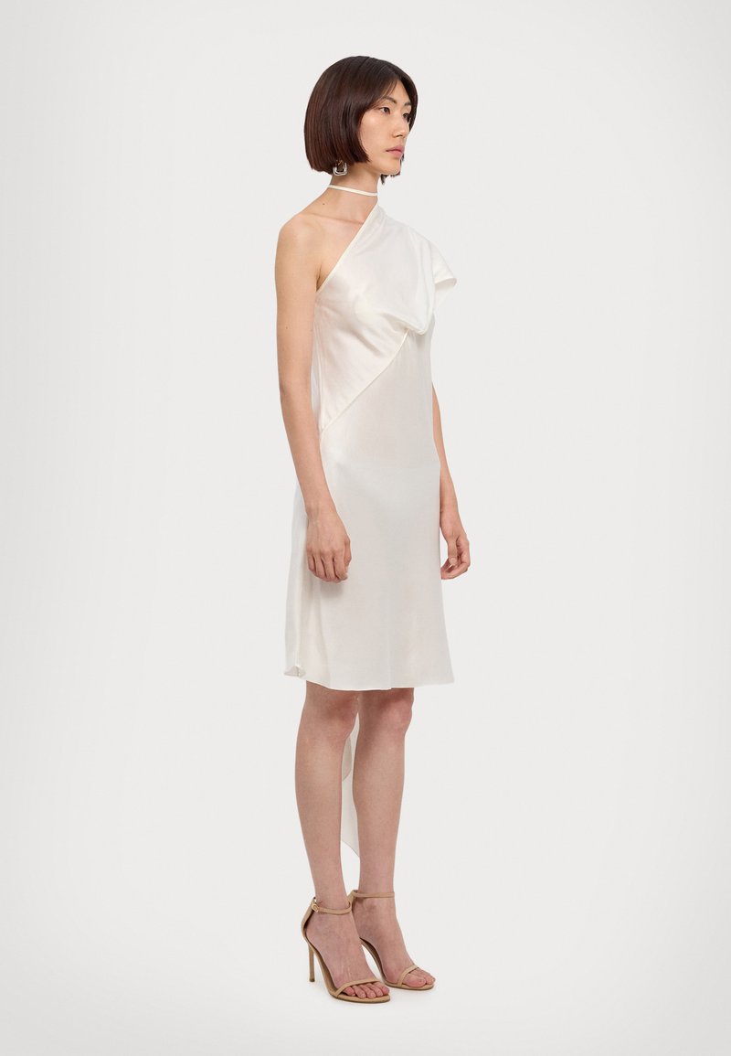 One-shoulder ivory dress with a draped overlay, made of smooth material. Model paired with nude heeled sandals, standing against a plain background.