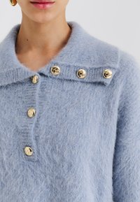 Ανοιχτό μπλε πλεκτό πουλόβερ με απαλή, αφράτη υφή. Διαθέτει σχεδιασμό με γιακά και τέσσερα χρυσά διακοσμητικά κουμπιά στο μπροστινό μέρος.
