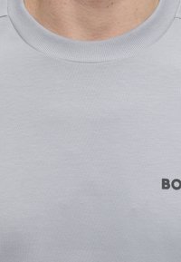 Gros plan d'une personne portant un sweat-shirt gris clair à col rond avec un texte noir partiel "BO" visible sur la poitrine.