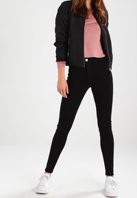 Černá bomber bunda přes růžový top, doplněná o černé džíny s vysokým pasem a bílé tenisky. Bunda má zapínání na zip a žebrované manžety.