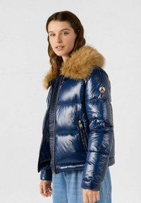 Marineblauwe donsjas met glanzende afwerking, voorzien van een afneembare nepbonten kraag, ritssluiting en zijzakken. Gladde textuur en doorgestikte design.