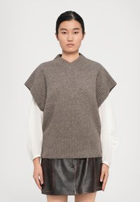 Frau trägt einen grauen ärmellosen Strickwesten über einer weißen Bluse mit Puffärmeln und einem schwarzen Lederrock, steht vor einem schlichten Hintergrund.