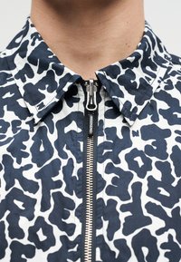 Γιακάς και φερμουάρ μαύρου και άσπρου μπουφάν με αφηρημένο μοτίβο και το κείμενο "WEEKEND" στο τραβηχτό του φερμουάρ, φοράει πρόσωπο με ορατό λαιμό.