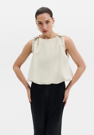 Donna con capelli pettinati all'indietro indossa un top beige senza maniche con dettagli a nodi sulle spalle e pantaloni neri a vita alta, mani sui fianchi.