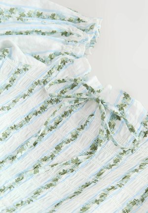 Fehér anyag függőleges kék csíkokkal és zöld virágmintákkal, ráncolt szakaszokkal és a nyakkivágásnál megkötött masnival.