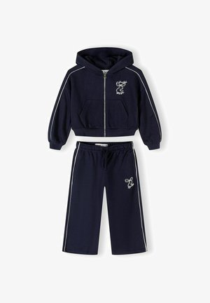 Sötétkék gyerek szabadidőruha fehér csíkozással, cipzáras kapucnis pulóverrel, elülső zsebekkel, gumírozott derekú nadrággal, valamint a mellkason és a lábszáron kis fehér masni hímzéssel.