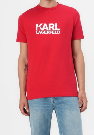 Persona que lleva una camiseta de manga corta roja con texto blanco "KARL LAGERFELD" y vaqueros azul claro sobre un fondo liso.