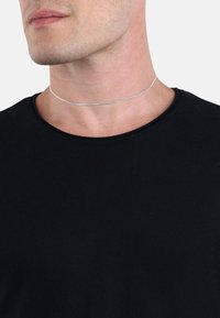 Collana choker d'argento con finitura testurizzata, che si adagia strettamente attorno al collo, abbinata a una maglietta nera a collo rotondo.