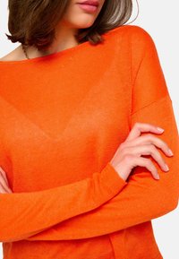 Pull en tricot orange avec un large col et des manches longues. Texture douce et poignets côtelés, montrant des bras croisés devant.
