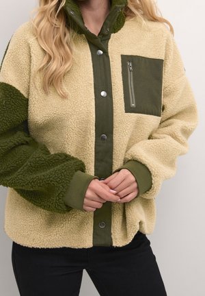 Femme portant une veste sherpa polaire beige et vert foncé avec boutons-pression et une poche poitrine zippée, mains librement croisées à la taille.