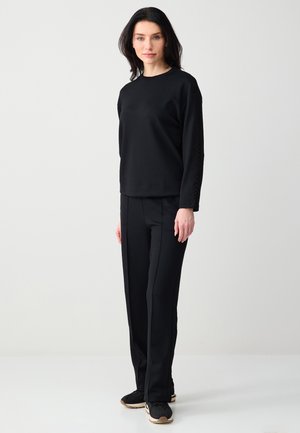 Kobieta stoi, ubrana w czarną bluzkę z długim rękawem i czarne spodnie proste, w czarno-beżowych tenisówkach, na tle jasnego, jednolitego tła.