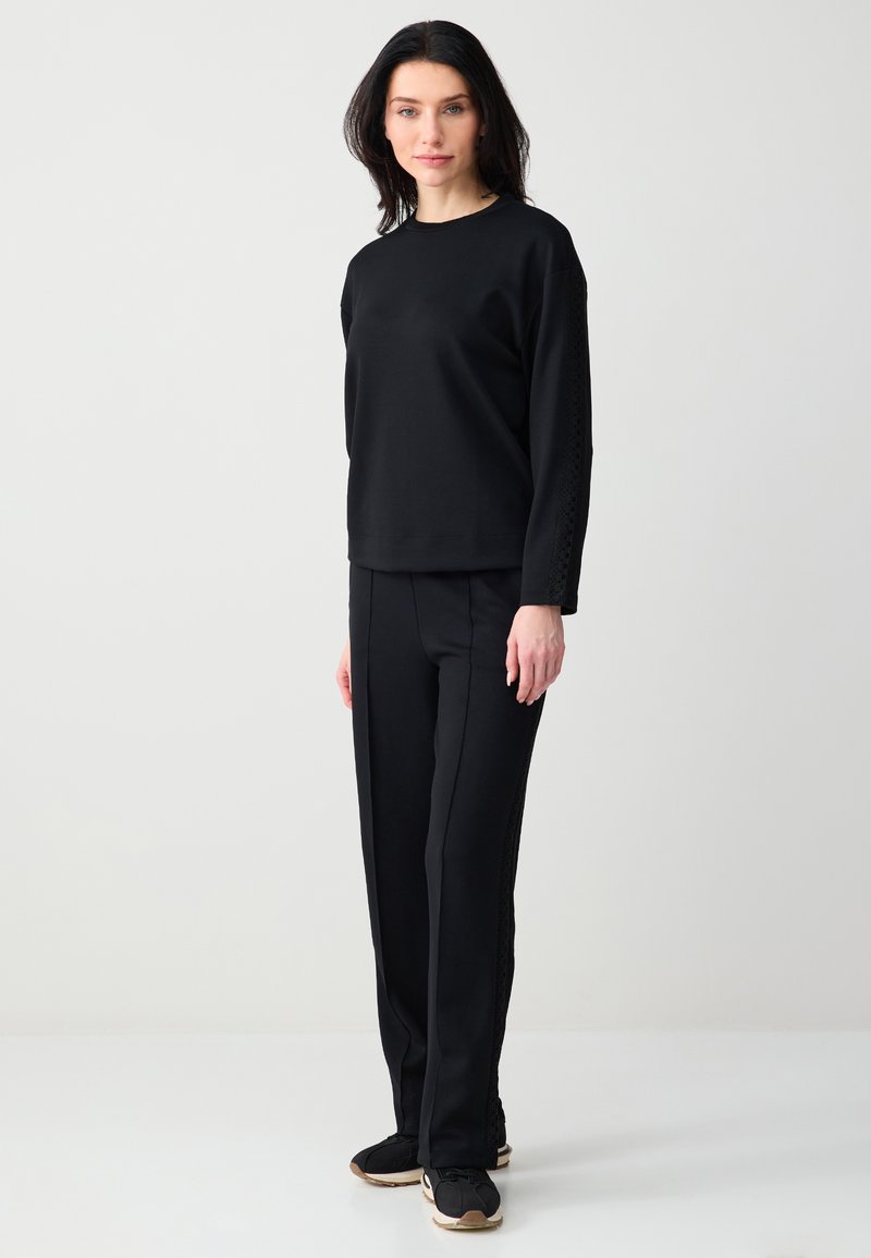 Femme debout portant un haut à manches longues noir et un pantalon droit noir avec des baskets noires et beiges, sur un fond clair uni.