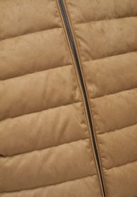 Chaleco acolchado color beige con textura de paja vertical, que presenta una cremallera negra central y material suave similar al ante.