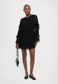 Black chiffon dress with long, ruffled sleeves, a round neckline, and an elasticized waist. Model holds a small black handbag.