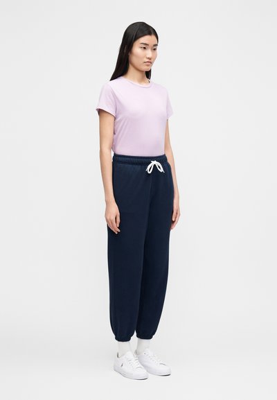 Ung kvinne som står, iført en lys lilla t-skjorte, marineblå joggebukser med hvit snøring, hvite sokker og hvite joggesko.