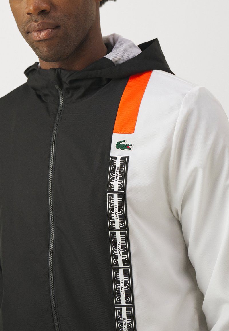 Uomo che indossa una giacca zip-up nera, bianca e arancione con cappuccio e branding verticale "Lacoste Sport" con logo coccodrillo verde sul petto.