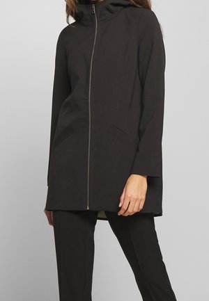 Femme portant une veste noire zippée avec un col montant et un pantalon noir assorti sur un fond gris uni.