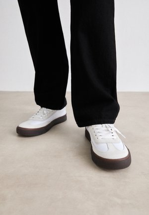 Personne portant des baskets blanches avec des semelles marron et un pantalon large noir, debout sur un sol beige.