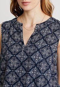 Donna che indossa un top senza maniche blu navy con un ornato motivo floreale bianco e una sottile collana d'oro.