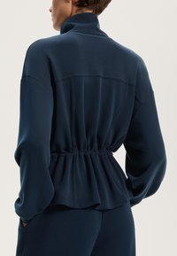 Tamno plava majica dugih rukava s visokim ovratnikom, skupljenim po struku i opuštenim krojem. Glatka tkanina s suptilnim sjajem i minimalnim detaljima šivanja.